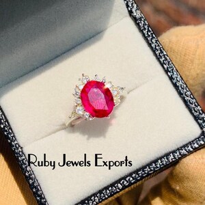 Peut inclure: Une bague en argent avec une grande pierre de rubis rouge ovale entourée de petites pierres blanches. La bague est dans un écrin à bague noir avec le texte "Ruby Jewels Exports".