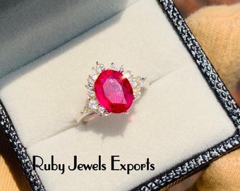 May include: A silver ring with a large oval-shaped red ruby gemstone surrounded by smaller white gemstones. The ring is in a black ring box with the text "Ruby Jewels Exports".
