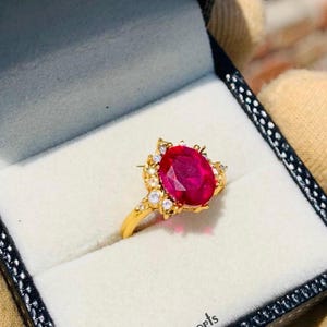 Peut inclure: Une bague en or avec une grande pierre de rubis ovale entourée de petites pierres blanches. La bague est dans un écrin à bague noir avec les mots "Ruby Jewels Exports" imprimés à l'intérieur.