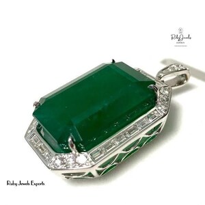 May include: A large emerald green gemstone pendant set in a silver frame with a diamond halo. The pendant is on a white background.