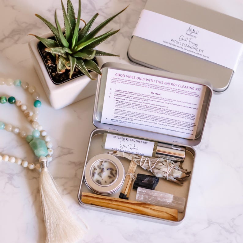 May include: A silver metal box with a white label that reads "Good Vibes Only With This Energy Clearing Kit". The box contains a white candle, a small bottle of spray, a bundle of sage, a small piece of black charcoal, a wooden stick, and a clear crystal.
