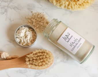 May include: A glass jar of Milky Way Cleansing Grains by Aligned Apothecary, a skincare product. The jar is on a white marble surface with a wooden brush and a natural sponge.