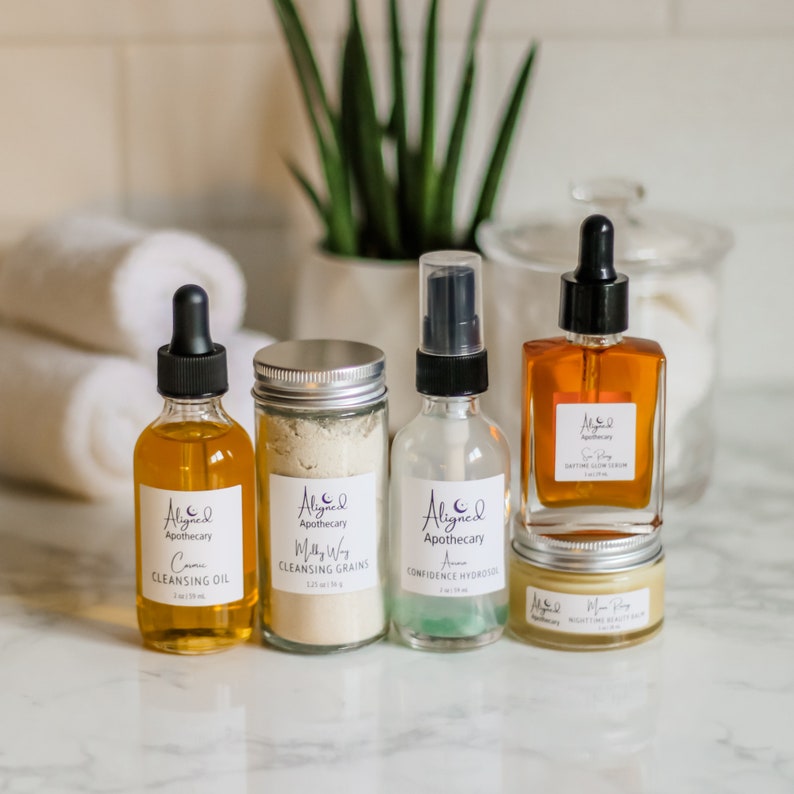 May include: A set of five skincare products in glass bottles and jars with silver lids. The products are labeled "Aligned Apothecary" and include a cleansing oil, cleansing grains, a serum, a daytime glow lotion, and a nighttime beauty balm.