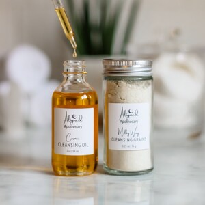 May include: Two glass bottles with white labels. The bottle on the left is filled with a yellow liquid and labeled "Aligned Apothecary Cosmic Cleansing Oil 2 oz | 59 ml". The bottle on the right is filled with a white powder and labeled "Aligned Apothecary Milky Way Cleansing Grains 1.25 oz | 36 g".