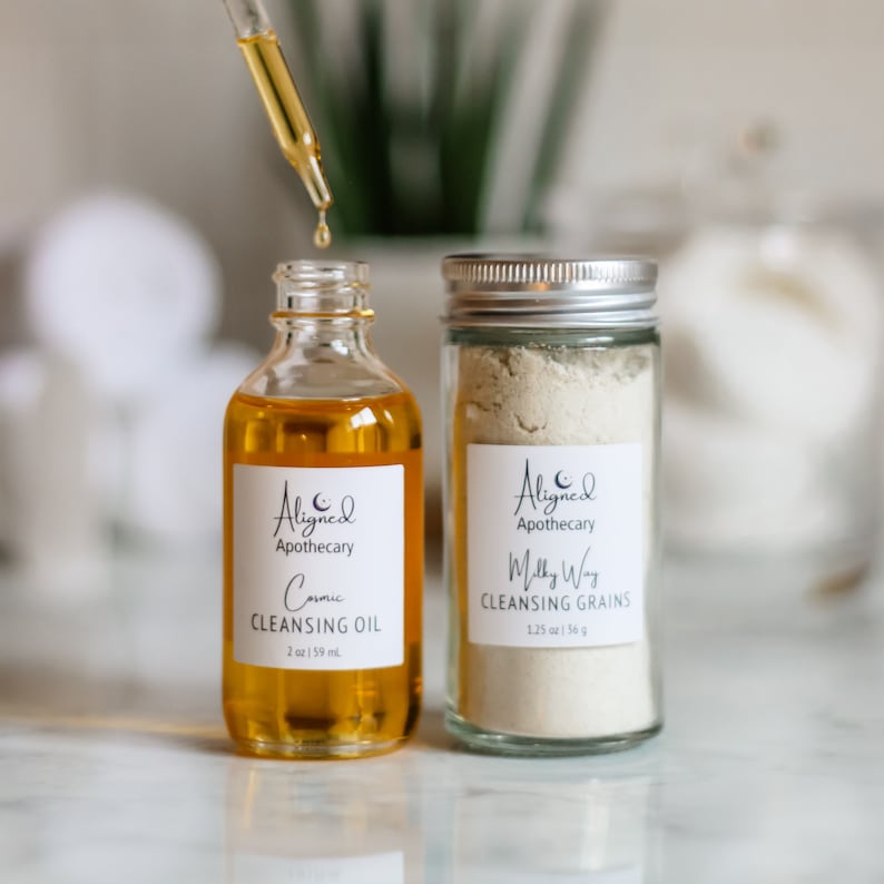 May include: Two glass bottles with white labels. The bottle on the left is filled with a yellow liquid and labeled "Aligned Apothecary Cosmic Cleansing Oil 2 oz | 59 ml". The bottle on the right is filled with a white powder and labeled "Aligned Apothecary Milky Way Cleansing Grains 1.25 oz | 36 g".
