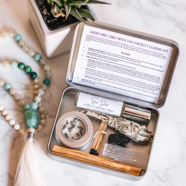 May include: A silver metal box with a white label that reads "Good Vibes Only With This Energy Clearing Kit" and instructions for a ritual. The box contains a small bottle of aura cleansing spray, a white candle, a sage smudge stick, a black crystal, a wooden stick of incense, and matches.