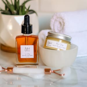 May include: Two skincare products, a clear glass bottle with a dropper labeled "Sun Ray Daytime Glow Serum 1 fl oz / 29 ml" and a white jar labeled "Moon Ray Nighttime Beauty Balm 1 oz / 28 ml", both with the brand name "Aligned Apothecary" on a white marble surface.