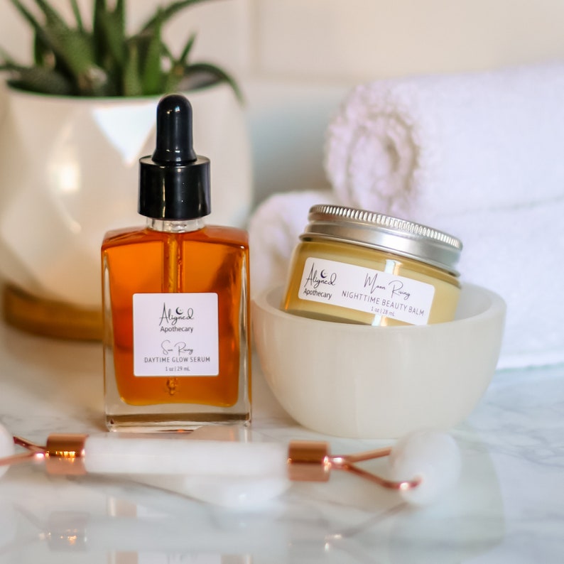 May include: Two skincare products, a clear glass bottle with a dropper labeled "Sun Ray Daytime Glow Serum 1 fl oz / 29 ml" and a white jar labeled "Moon Ray Nighttime Beauty Balm 1 oz / 28 ml", both with the brand name "Aligned Apothecary" on a white marble surface.