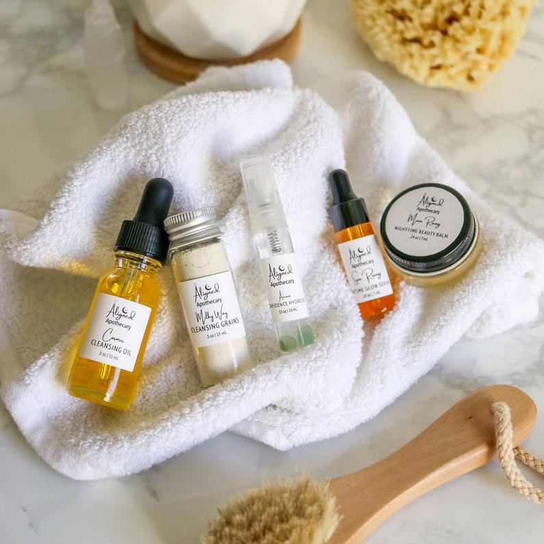 May include: A set of five skincare products in glass bottles and jars with white labels and gold lettering. The products include a cleansing oil, cleansing grains, a nighttime beauty balm, a serum, and a face mist. The products are arranged on a white towel with a wooden brush and a loofah sponge in the background.