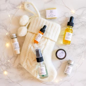 May include: A white knit Christmas stocking filled with skincare products, including a facial bar soap, cleansing oil, and face mask. The products are arranged on a marble surface with string lights.