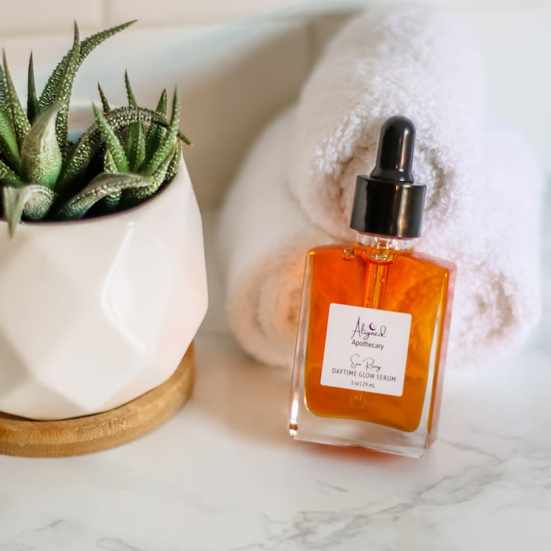 May include: A clear glass bottle with a dropper filled with a golden liquid. The bottle is labeled "Aligned Apothecary Sun Rose Daytime Glow Serum 1 oz (29 ml)".