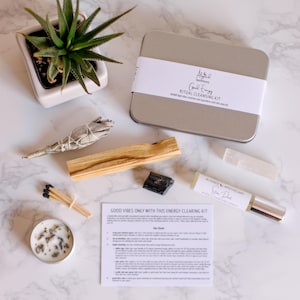 May include: A silver tin box with a white label that reads "Good Energy Ritual Cleansing Kit" and contains a variety of items for energy clearing, including sage, palo santo, a crystal, matches, a candle, and a bottle of essential oil.