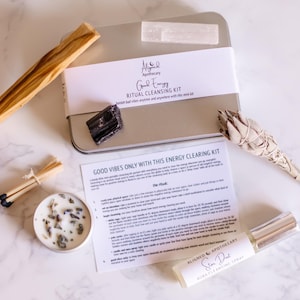 May include: A silver metal box with a white label that reads "Good Energy Ritual Cleansing Kit" and a white label that reads "Aligned Apothecary". The box contains a white sage smudge stick, a black obsidian crystal, a clear quartz crystal, and a small white candle. The box is sitting on a white marble surface with a white and brown wooden stick, a pack of matches, and a small bottle of "Star Dust Aura Cleansing Spray" by Aligned Apothecary.