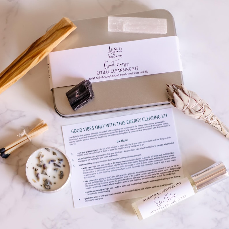 May include: A silver metal box with a white label that reads "Good Energy Ritual Cleansing Kit" and a white label that reads "Aligned Apothecary". The box contains a white sage smudge stick, a black obsidian crystal, a clear quartz crystal, and a small white candle. The box is sitting on a white marble surface with a white and brown wooden stick, a pack of matches, and a small bottle of "Star Dust Aura Cleansing Spray" by Aligned Apothecary.