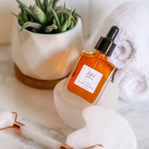 May include: A glass bottle of amber-colored serum with a black dropper top. The bottle is labeled "Aligned Apothecary Sun Body Daytime Glow Serum 1 fl oz/29 ml".