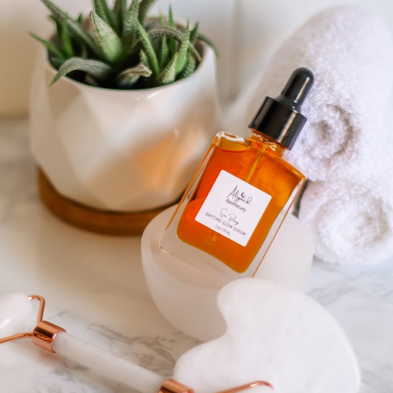 May include: A glass bottle of amber-colored serum with a black dropper top. The bottle is labeled "Aligned Apothecary Sun Body Daytime Glow Serum 1 fl oz/29 ml".