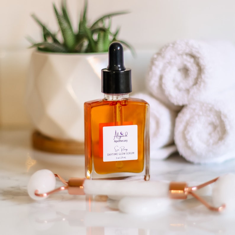 May include: A glass bottle of serum with a dropper, labeled "Aligned Apothecary Sun Ray Daytime Glow Serum 1 fl oz / 29 ml". The bottle is sitting on a white marble surface with two white towels and a white jade roller.