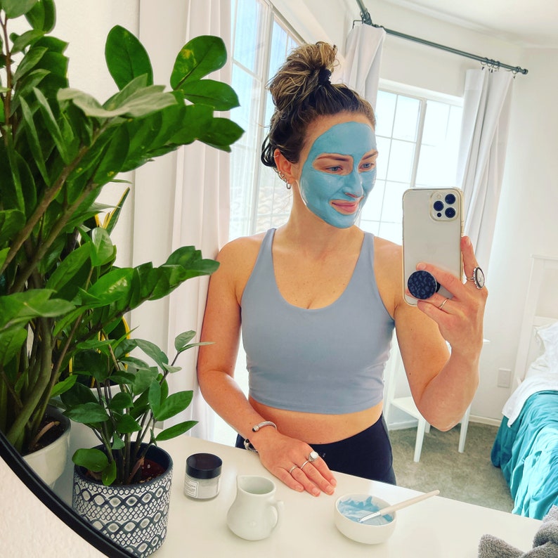 May include: A woman wearing a light blue sports bra and a blue face mask is taking a selfie with a smartphone. She is holding the phone in her right hand and has a ring on her left hand. There is a small bowl with a white substance in it and a white cup on the counter.