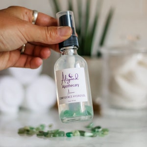 May include: A clear glass bottle with a black spray top and a white label that reads "Aligned Apothecary Aura Confidence Hydrosol 2 oz / 59 ml". The bottle is filled with a clear liquid and contains small green crystals.