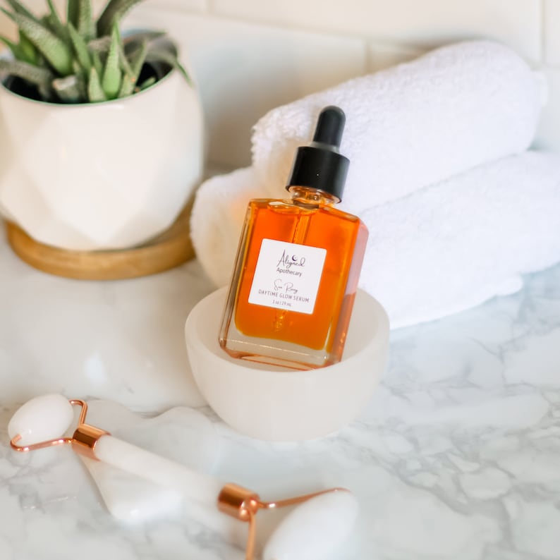 May include: A glass bottle of amber-colored serum with a dropper top. The bottle is labeled "Alche. Apothecary" and "Sun Glow Serum Daytime Glow Serum 1 fl oz (30 ml)". The bottle is sitting on a white pedestal.