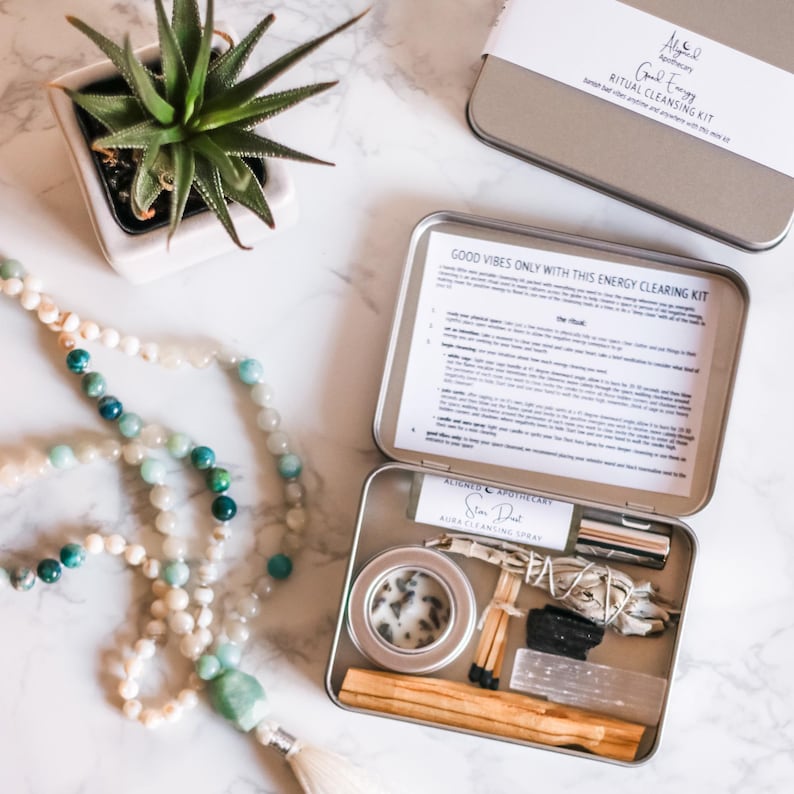 May include: A silver metal box with a white label that reads "Good Energy Ritual Cleansing Kit". The box is open and contains a variety of items, including a small white candle, a bundle of sage, a small bottle of spray, a pack of matches, and a wooden stick of incense. The box is sitting on a white marble surface.