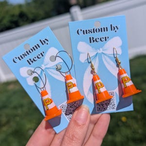 May include: Two pairs of earrings featuring miniature orange and white traffic cones with a yellow letter P printed on the side. The earrings are attached to light blue display cards with the text Custom by Beer and a white bow graphic.