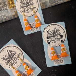 May include: Three pairs of miniature orange and white traffic cone earrings, each featuring a yellow letter P. The earrings are displayed on light blue cards with a circular logo that reads The Crafted Chapter and a small QR code at the bottom.
