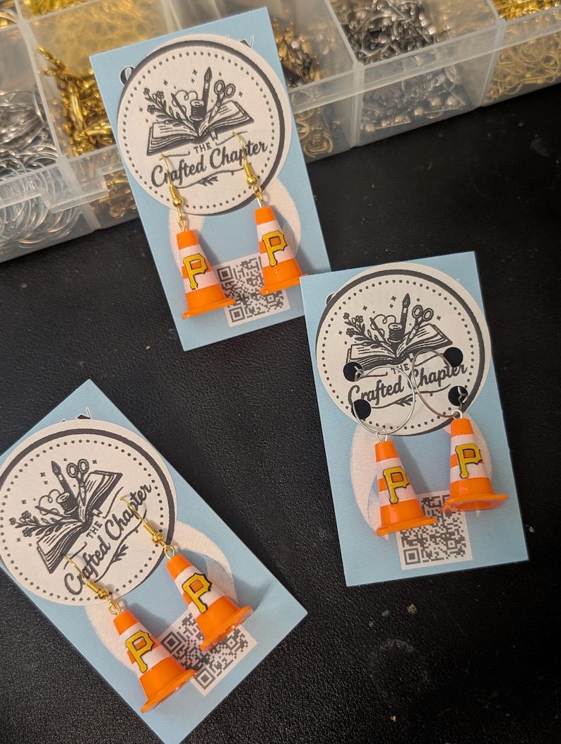 May include: Three pairs of miniature orange and white traffic cone earrings, each featuring a yellow letter P. The earrings are displayed on light blue cards with a circular logo that reads The Crafted Chapter and a small QR code at the bottom.