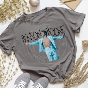 May include: Gray t-shirt featuring a graphic of Benson Boone in a light blue suit with the text "Benson Boone" in black. The shirt is made of a soft material and is suitable for casual wear.