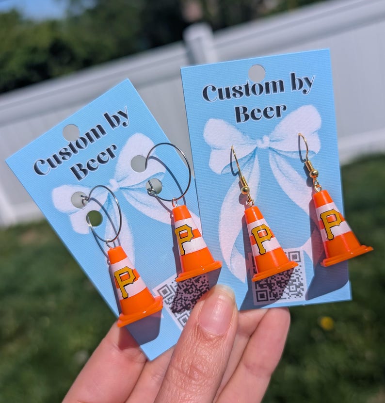 May include: Two pairs of earrings featuring miniature orange and white traffic cones with a yellow letter P printed on the side. The earrings are attached to light blue display cards with the text Custom by Beer and a white bow graphic.