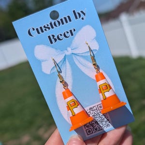 May include: A pair of dangle earrings featuring miniature orange and white traffic cones with a black letter P logo. The earrings are attached to a light blue display card with the text Custom by Beer and a white bow graphic printed on it.