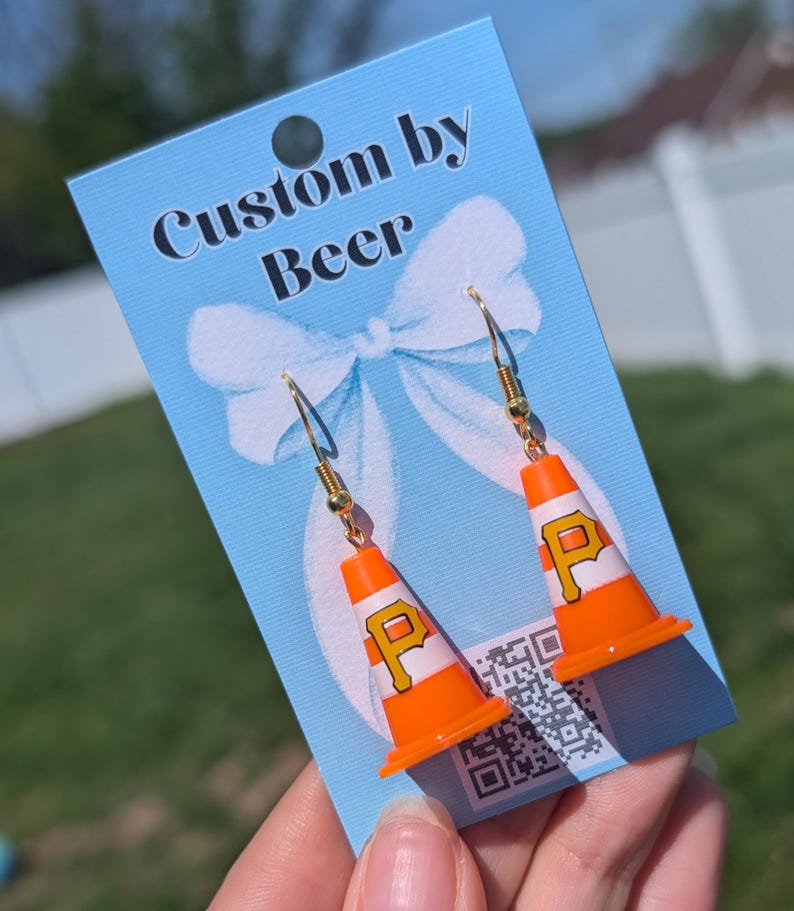 May include: A pair of dangle earrings featuring miniature orange and white traffic cones with a black letter P logo. The earrings are attached to a light blue display card with the text Custom by Beer and a white bow graphic printed on it.