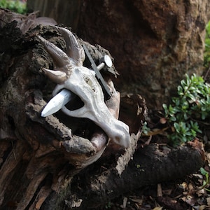 Könnte beinhalten: Eine weiße Keramik-Drachen-Skulptur mit einer silbernen Klinge durch den Kopf, die auf einem rauen, braunen Baumstamm ruht.