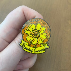 May include: A yellow and brown enamel pin featuring a stylized illustration of a Lanceleaf Coreopsis flower with the text "Lanceleaf Coreopsis" on a banner.