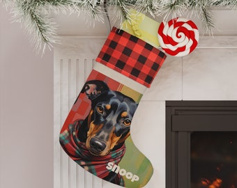 Op de afbeelding: Een rode en zwarte geruite kerstsok met een hondenkop erop. De hond draagt een rode en groene geruite sjaal en de sok heeft de tekst "Snoop" erop. Er zit een rode en witte pepermuntstok bovenop de sok.