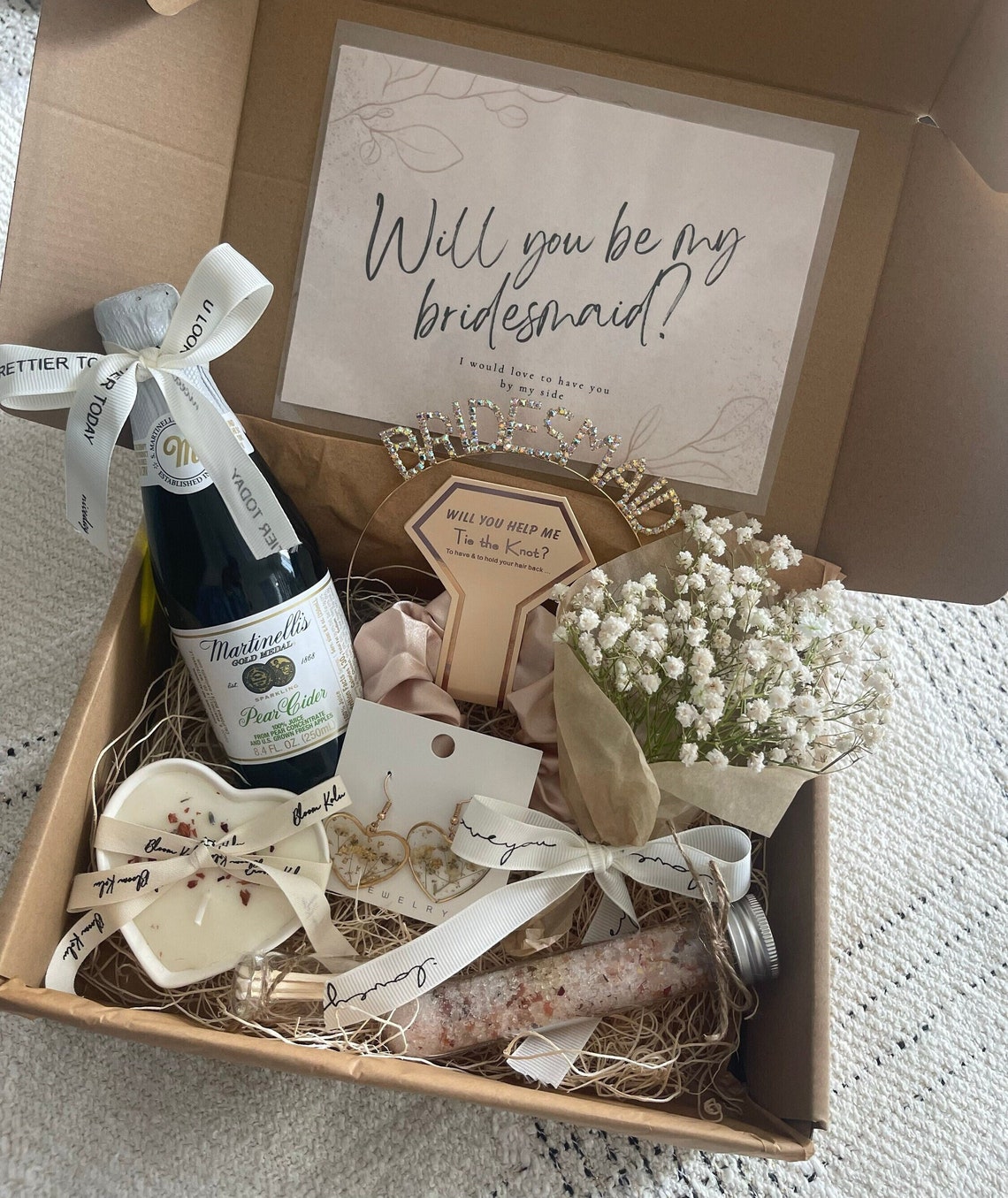 May include: A gift box filled with items for a bridesmaid proposal. The box includes a bottle of pear cider, a heart-shaped candle, a pair of earrings, a small bouquet of white flowers, a headband with the word "bridesmaid" on it, and a card that says "Will you be my bridesmaid?"