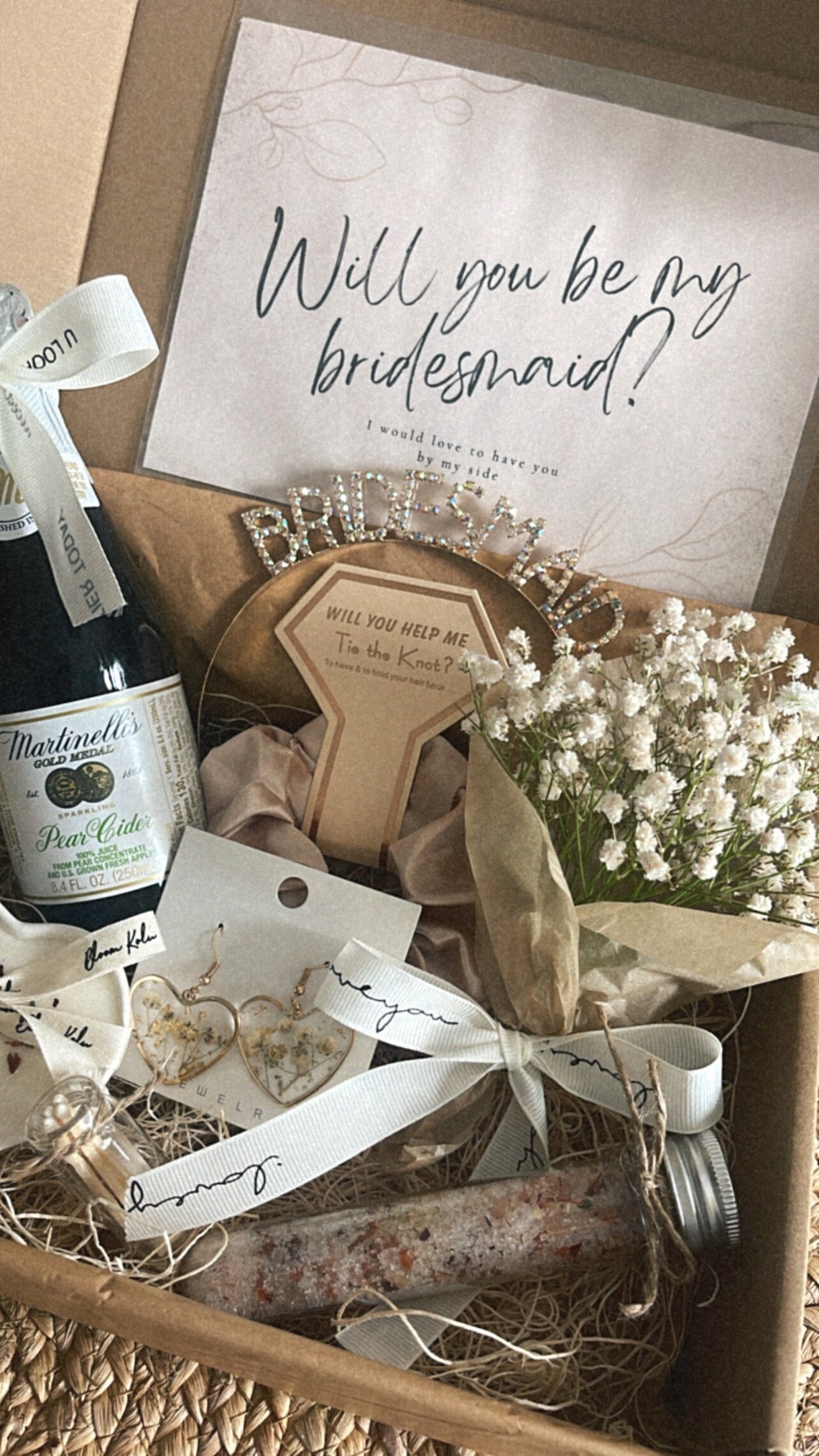 May include: A gift box filled with items for a bridesmaid proposal. The box includes a card that says "Will you be my bridesmaid?", a small bottle of pear cider, a headband, a small bouquet of white flowers, a pair of earrings, a small bottle of bath salts, and a wooden sign that says "Will you help me tie the knot?"