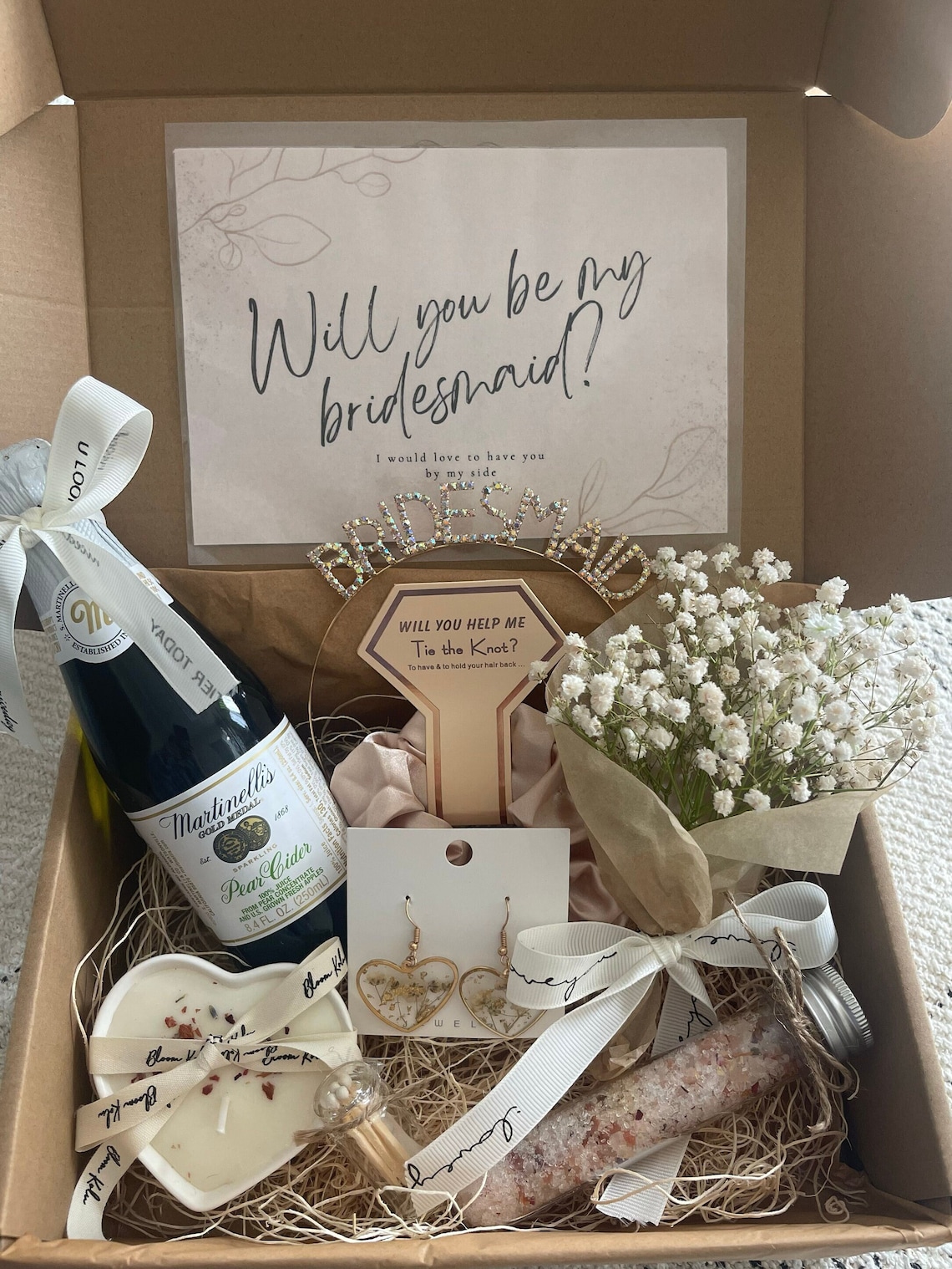 May include: A gift box filled with items for a bridesmaid. The box contains a bottle of pear cider, a bouquet of white flowers, a heart-shaped candle, a small bottle of bath salts, a pair of earrings, and a card that says "Will you be my bridesmaid?"