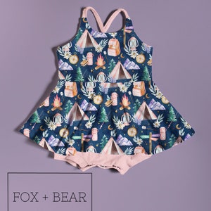 May include: A navy blue baby romper with a camping-themed pattern featuring tents, campfires, backpacks, and compasses. The romper has a pink ribbed bottom and straps. The text "FOX + BEAR Kids Co" is displayed in a box at the bottom.
