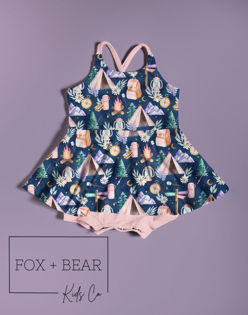 May include: A navy blue baby romper with a camping-themed pattern featuring tents, campfires, backpacks, and compasses. The romper has a pink ribbed bottom and straps. The text "FOX + BEAR Kids Co" is displayed in a box at the bottom.