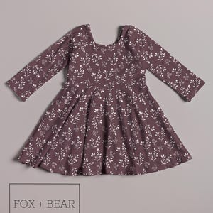 May include: A long-sleeved, plum-colored dress with a white leaf pattern. The dress has a scoop back and a flared skirt. The text "FOX + BEAR Kids Co" is in a white rectangular box at the bottom of the image.