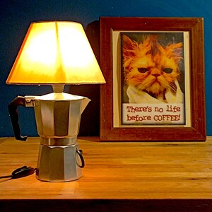 Può includere: Una caffettiera d'argento con un manico nero viene utilizzata come base per una lampada. Un paralume bianco con una luce gialla illumina la caffettiera. Una foto incorniciata di un gatto con il testo "There's no life before COFFEE!" è su un tavolo di legno.