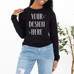 May include: Black long-sleeve shirt with the text "YOUR DESIGN HERE" in white. The shirt is paired with light blue ripped jeans. A woman is wearing the shirt. A plant is in the background.