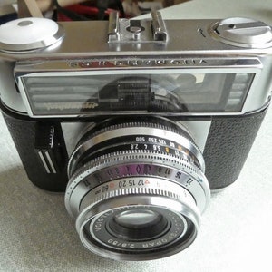 May include: A silver and black 35mm film camera with a silver lens. The lens has a black ring with white markings for aperture and shutter speed settings. The camera has a black leatherette covering and a silver top plate with the text "VITOMATIC II" printed on it.