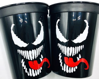 May include: Two black plastic cups with a white Venom character design. The character has a red tongue and white eyes.