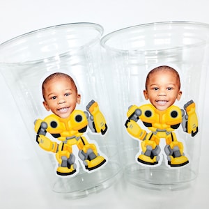 May include: Two clear plastic cups with yellow and black cartoon robot stickers featuring a child's face. The robot is in a superhero pose with arms raised.