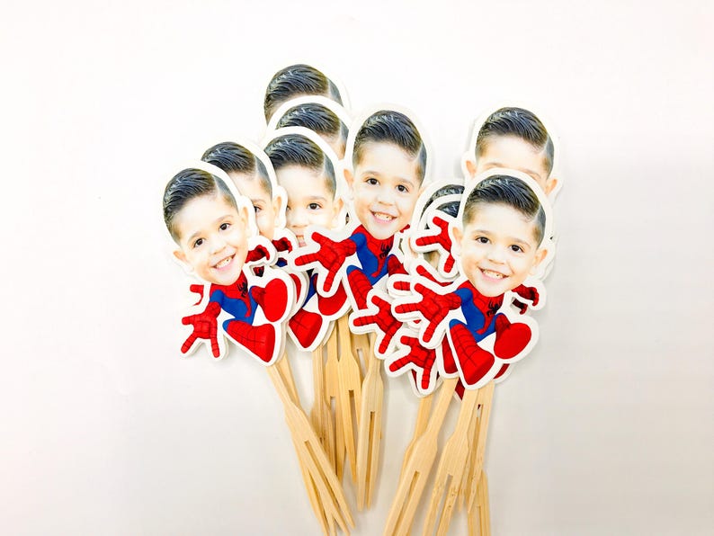 Puede incluir: Un conjunto de 10 adornos para cupcakes de Spiderman rojos y azules con una foto de la cara de un ni&ntilde;o en cada adorno. Los adornos est&aacute;n unidos a brochetas de madera.