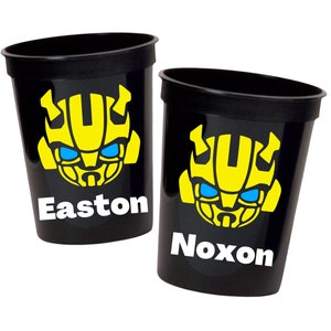 May include: Two black plastic cups with a yellow Bumblebee Transformer head graphic and blue eyes. The cups have the names Easton and Noxon printed on them.
