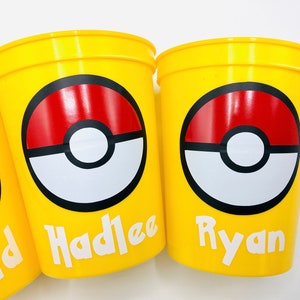 Pu&ograve; includere: Tre bicchieri di plastica gialli con un design Pokeball rosso, bianco e nero. I bicchieri hanno i nomi "David", "Hadlee" e "Ryan" stampati su di essi.