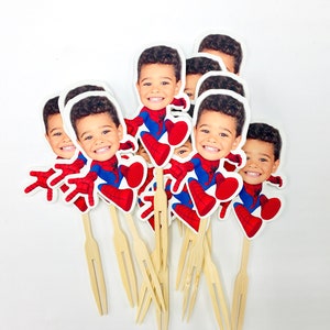 Puede incluir: Un conjunto de 8 adornos para cupcakes con la cara de un ni&ntilde;o con un disfraz de Spiderman. Los adornos est&aacute;n impresos en cartulina blanca y unidos a brochetas de madera.